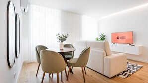 A modern living room with a sofa, two chairs, a dining table with a vase of flowers, a TV on a stand, and a large window with sheer curtains. - GuestReady - A marvellous city retreat (Porto)