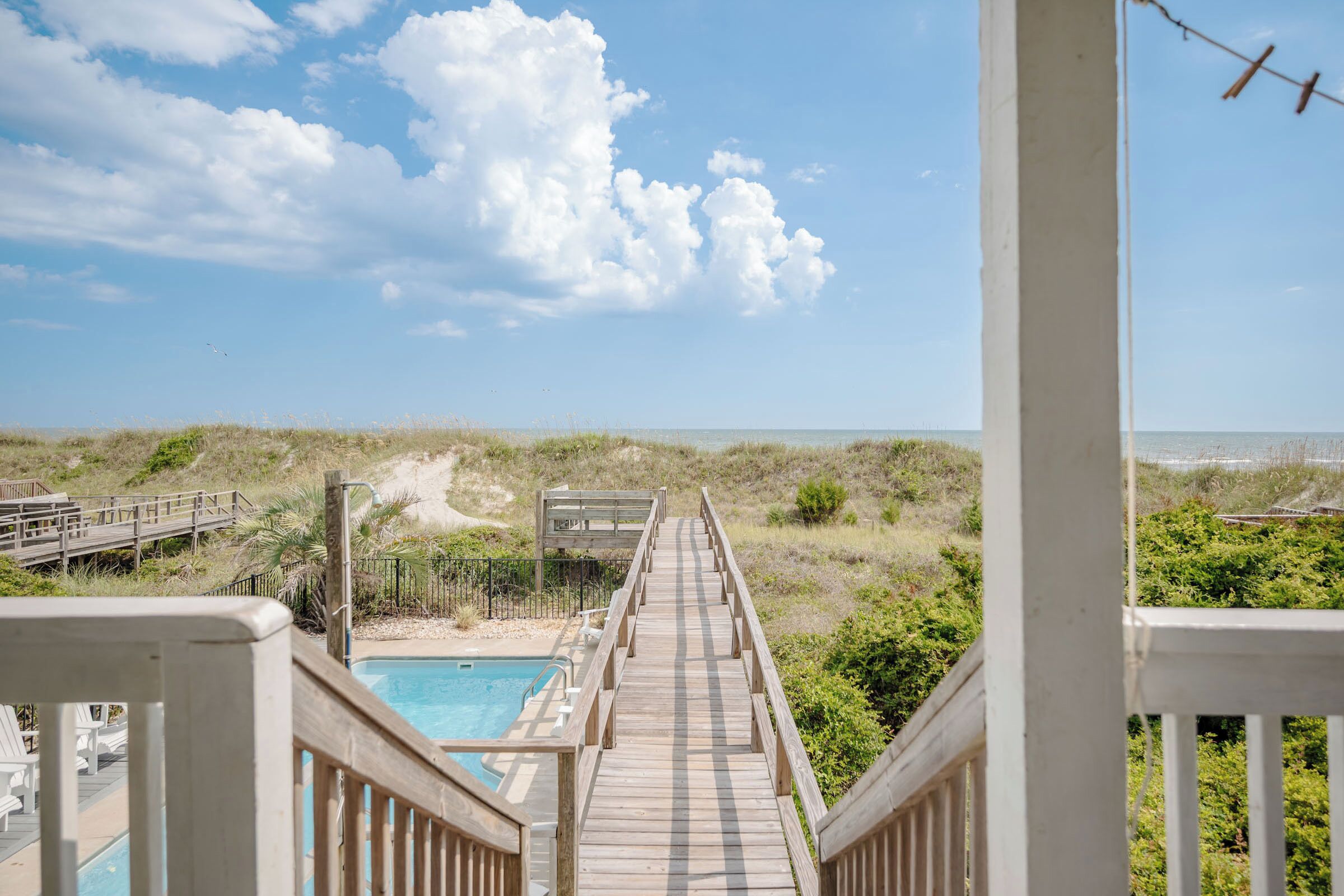 A wooden deck leading to a pool with a view of the beach and dunes.