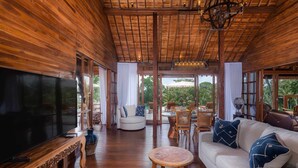 A spacious living area with a large flat-screen TV, wooden furniture, and large windows offering a view of the outdoors. - 1 BR Cozy Villa With Breathtaking Rice Paddy View (Bali)
