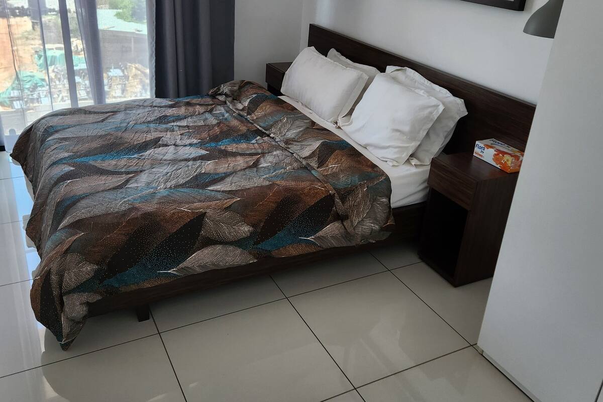 Una cama bien hecha con una colcha estampada, una mesita de noche con una caja y un cuadro en la pared.