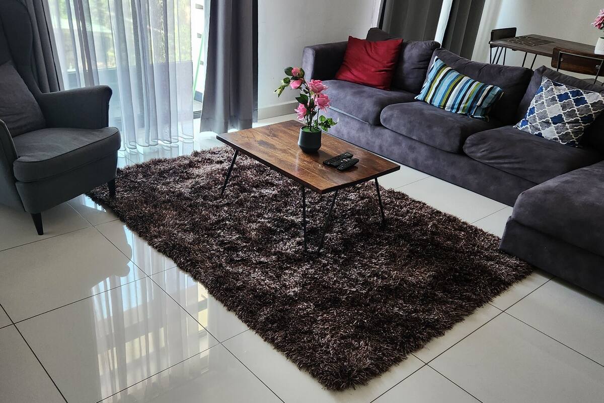 Una sala moderna con una alfombra mullida oscura, una mesa de centro de madera con un florero, un sofá gris con cojines estampados y un sillón gris.