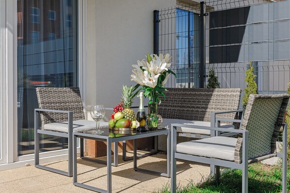 Relax in a serene outdoor space with a glass table and wicker chairs, surrounded by lush greenery and a beautiful flower arrangement.