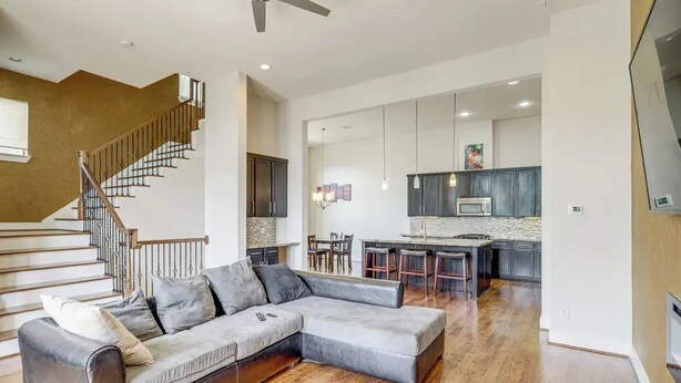 A spacious living area with a large grey sectional sofa, a wooden staircase, and a modern kitchen with dark cabinets and a microwave. - Houston Home w/ Rooftop Patio - 3 Mi to Downtown!
 (Houston)
