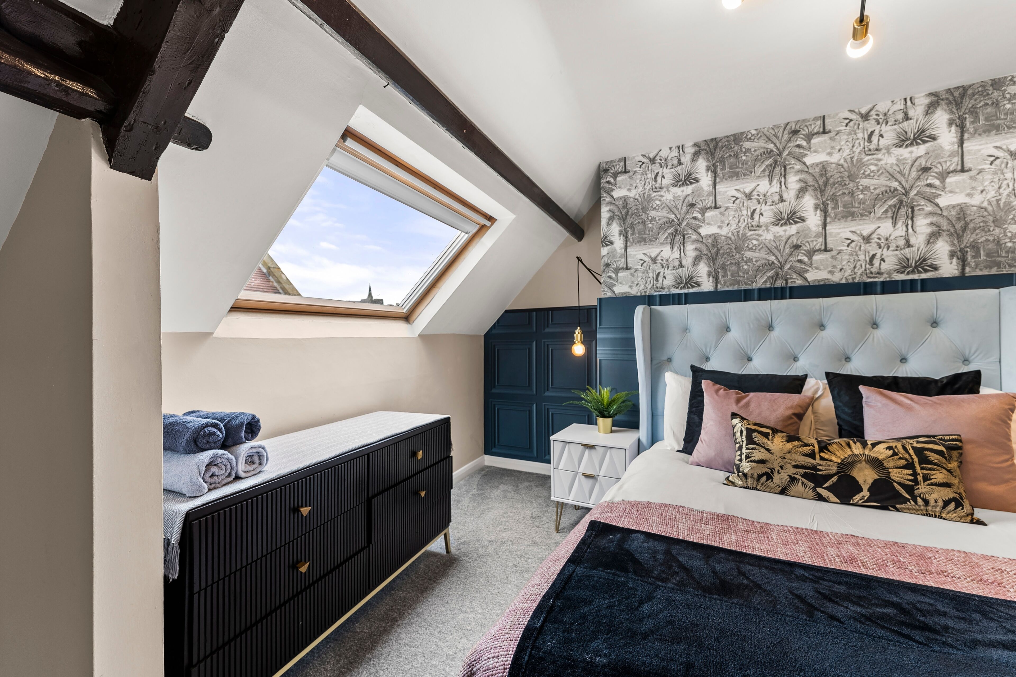 A bedroom with a skylight, a bed with multiple pillows, a bedside table with a plant, and a dark-colored dresser.