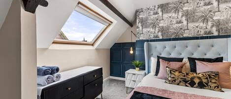 A bedroom with a skylight, a bed with multiple pillows, a bedside table with a plant, and a dark-colored dresser.