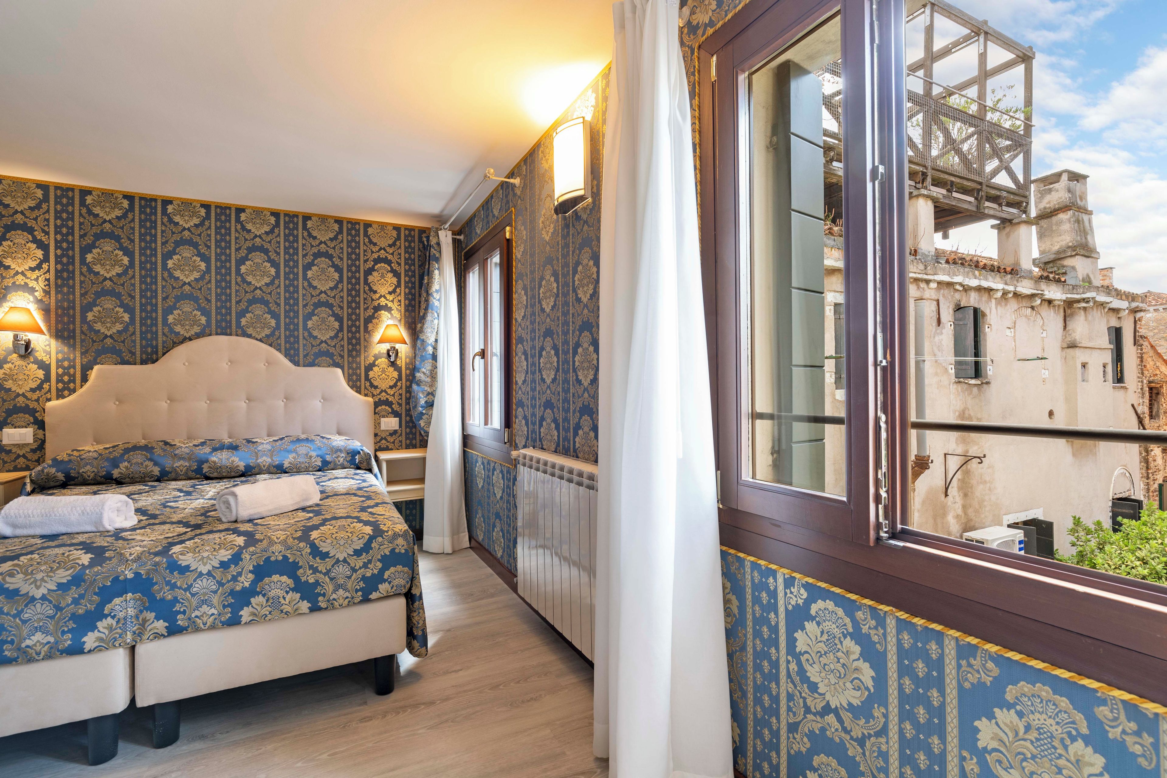 A bedroom with a large bed, a window with a view of a building, and patterned wallpaper.