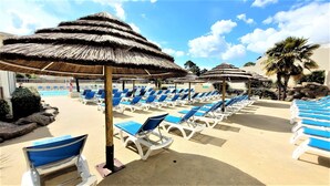 A poolside area with sun loungers and thatched umbrellas, surrounded by palm trees and rocks. - Mobil home 3 chambres (Saint-Jean-de-Monts)