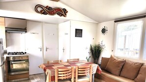 A compact kitchen with a stove, oven, and cabinets. There is a dining table with chairs and a sofa in the living area. - Mobil home 3 chambres (Saint-Jean-de-Monts)