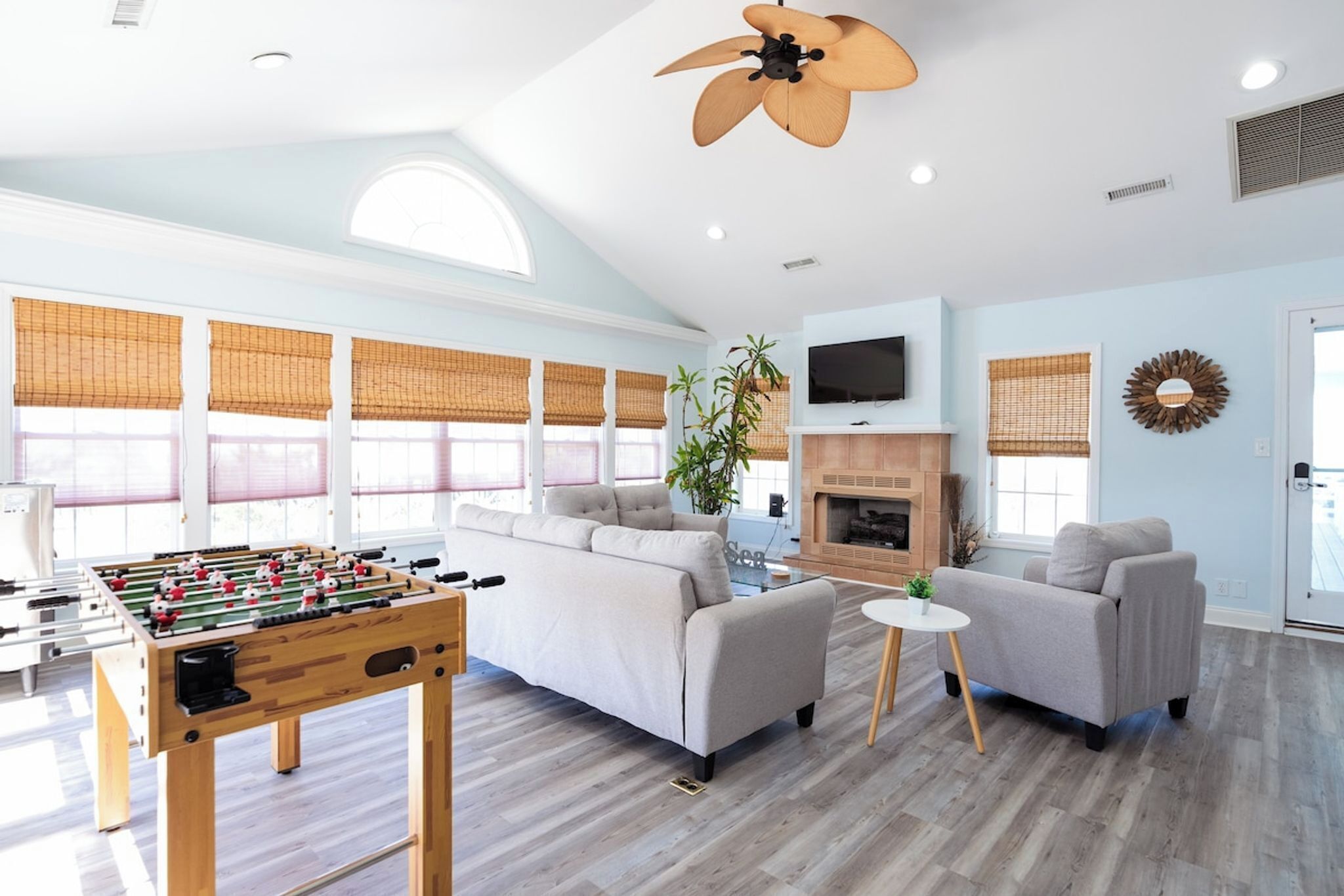 A spacious living room with a foosball table, sofas, a fireplace, and large windows with blinds.