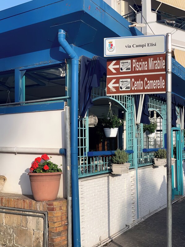 Un bâtiment à la façade bleue et blanche, une enseigne indiquant les directions vers « Piscina Mirabile » et « Cento Camerelle », et un pot de fleurs orné de fleurs rouges.