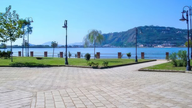 Un chemin pavé bordé de lampadaires, une zone herbeuse et une vue sur un lac avec des collines en arrière-plan.