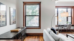 A modern living room with a wooden floor, a grey sofa, a desk with a lamp, and a large window with a view of trees and buildings.