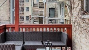 A covered outdoor seating area with wicker furniture and a glass table, overlooking a row of multi-story buildings.