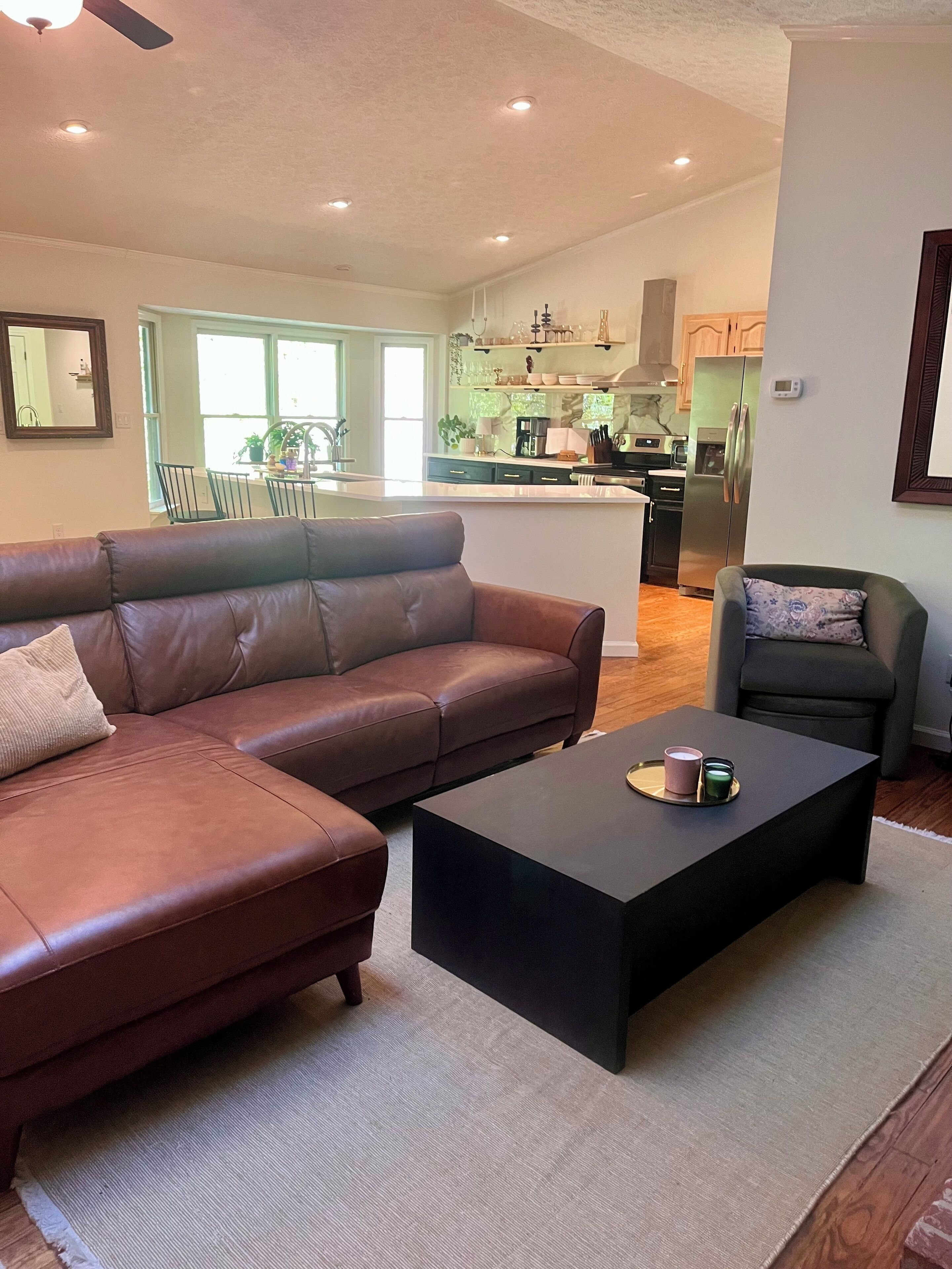 Een moderne woonkamer met een bruine leren bank, een zwarte salontafel en een grijze fauteuil. In de achtergrond is een keuken met een roestvrijstalen koelkast en een eethoek met een tafel en stoelen te zien.