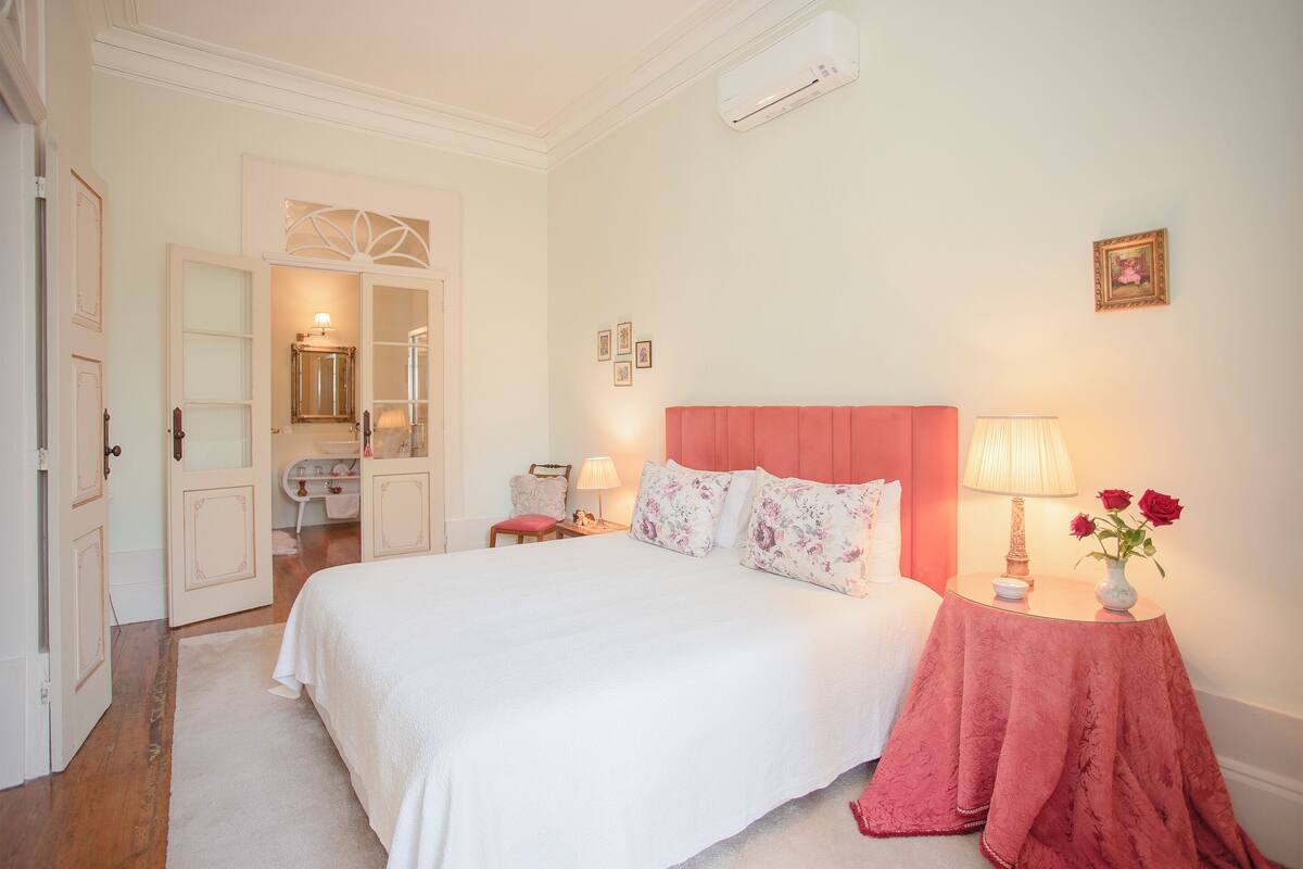 A bedroom with a large bed, a vanity with a mirror, a red headboard, and a bedside table with a lamp and flowers.