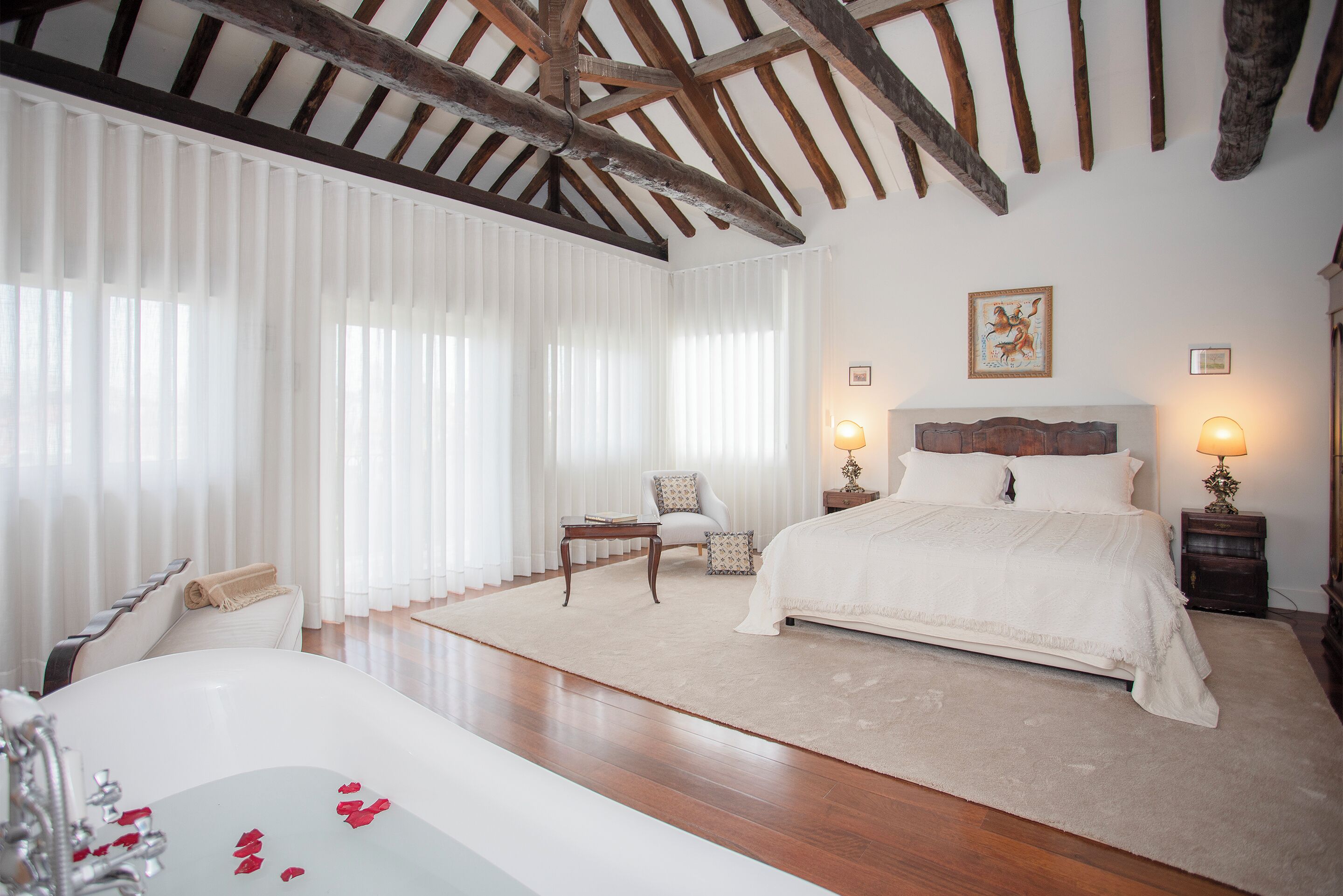 A bedroom with a large bed, a sitting area with a chair, a small table, and a bathtub with rose petals.