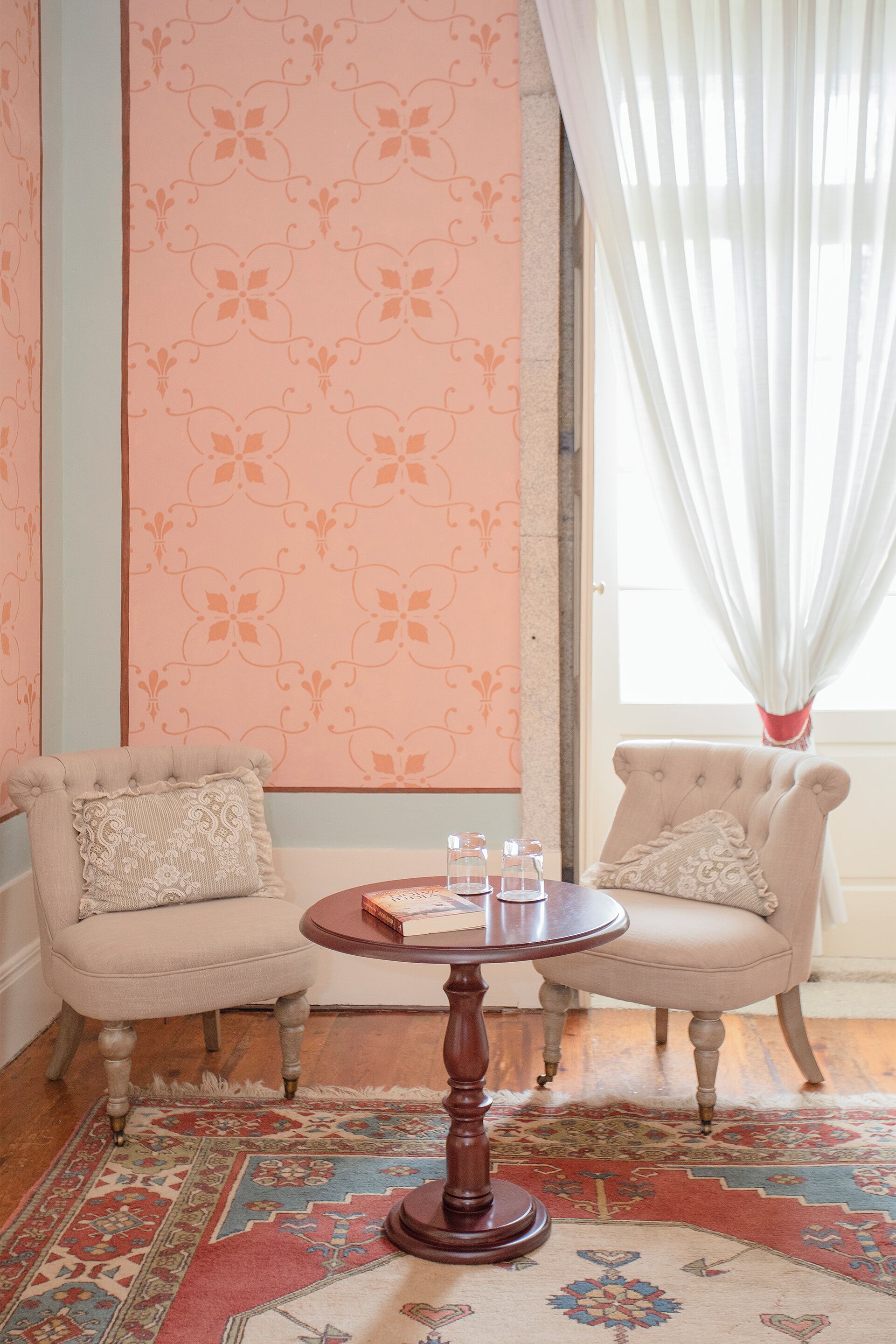 A room with two upholstered chairs, a round wooden table with a book and glasses, a patterned rug, and a window with sheer curtains.