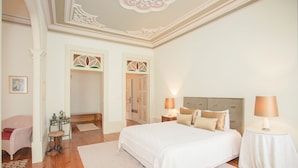 Une chambre à coucher avec un grand lit, deux tables de chevet avec des lampes, une petite table avec une chaise et une porte donnant sur une autre pièce.