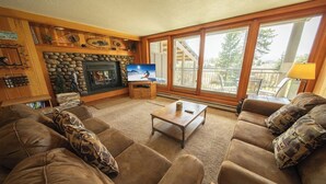 A cozy living room with a stone fireplace, a television, and large windows offering a view of the outdoors. - Family Fav, Indoor Pool/Hot Tubs, Shuttle (Keystone)