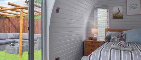 A bedroom with a large window, a bed with striped bedding, a nightstand with a lamp, and a view of a wooden deck and outdoor furniture.