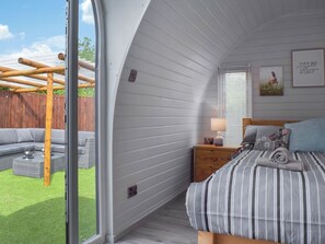 A bedroom with a large window, a bed with striped bedding, a nightstand with a lamp, and a view of a wooden deck and outdoor furniture. - Laurel Pod - UK47203 (Lathom, near Southport)