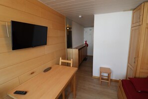 A compact room with a wooden table, a single chair, a small stool, a bed with red bedding, and a television mounted on the wall. - 10m from resort center, 3rd floor, tv, ski locker, 15m², Tignes (Tignes)