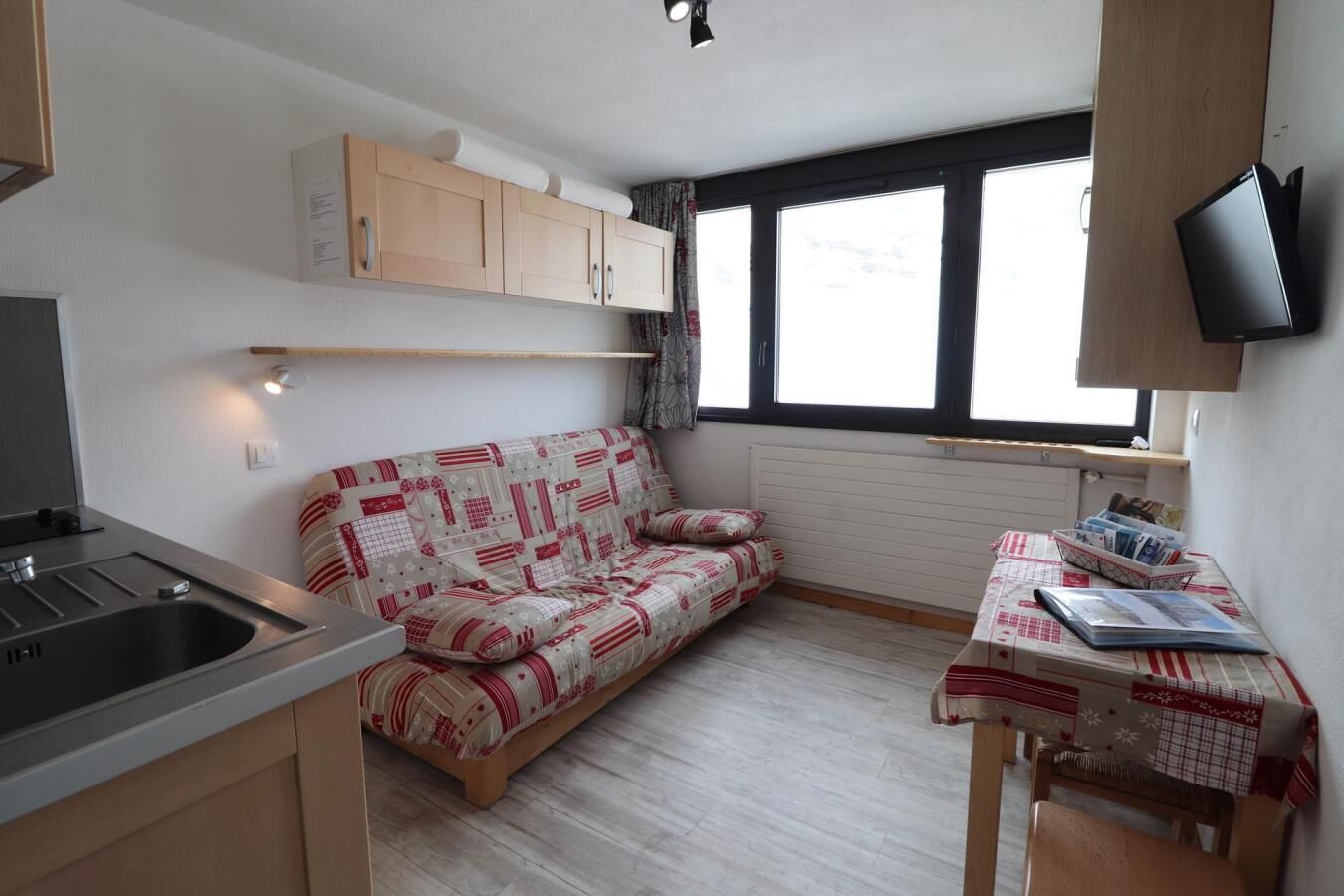 Eine kompakte Küchenzeile mit Spüle und Herd, ein Sofa mit gemustertem Bezug, ein kleiner Tisch mit Zeitschriften und ein Fenster mit Vorhängen.