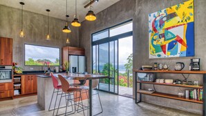 A modern kitchen with a large island, bar stools, and a view of greenery through large windows.