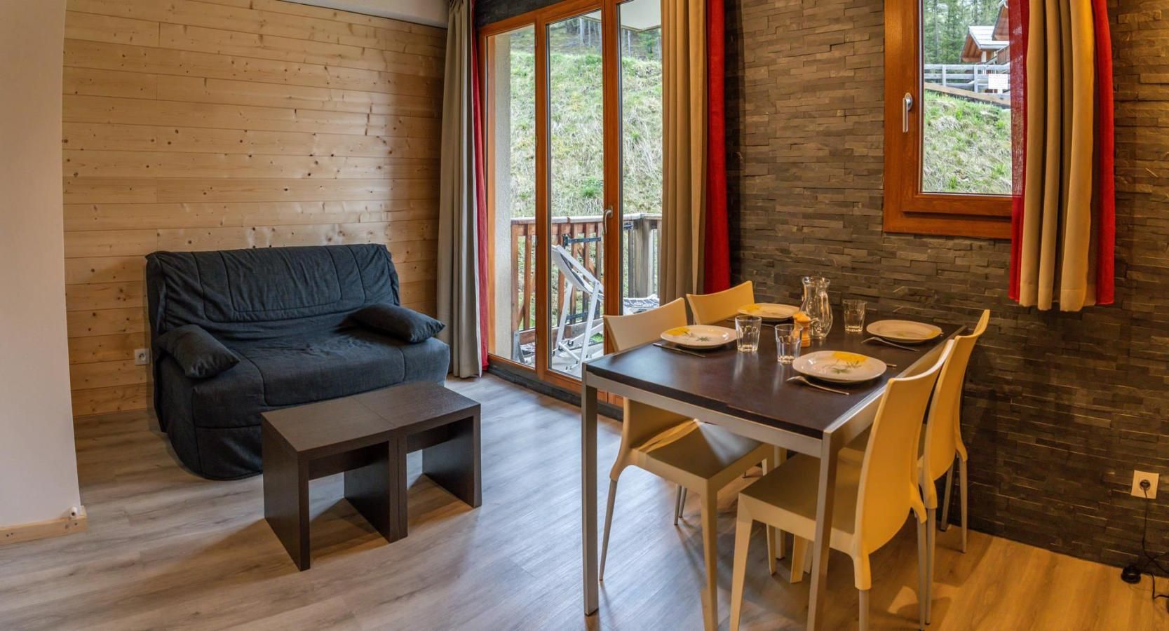 Ein Zimmer mit Holzboden, einem dunklen Sofa, einem kleinen Tisch und einer Essecke mit Stühlen und einem Tisch für zwei Personen.