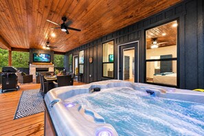 A hot tub in a covered outdoor area with a wooden deck, a fireplace, and a television mounted on the wall.