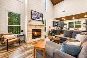 A modern living room with a fireplace, a sofa, a coffee table, and a kitchen area with cabinets and a dining table. - Family Retreat | Hot Tub | Arcade Games & Playset (Broken Bow)