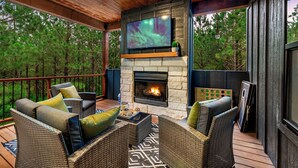 A covered deck with wicker furniture, a stone fireplace, and a TV mounted above it. - Family Retreat | Perfect Location | Hot Tub (Broken Bow)
