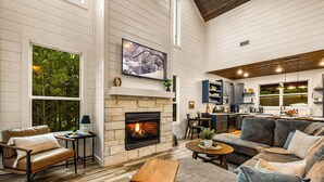 A spacious living room with a fireplace, a large window, and a kitchen area. - Family Retreat | Perfect Location | Hot Tub (Broken Bow)
