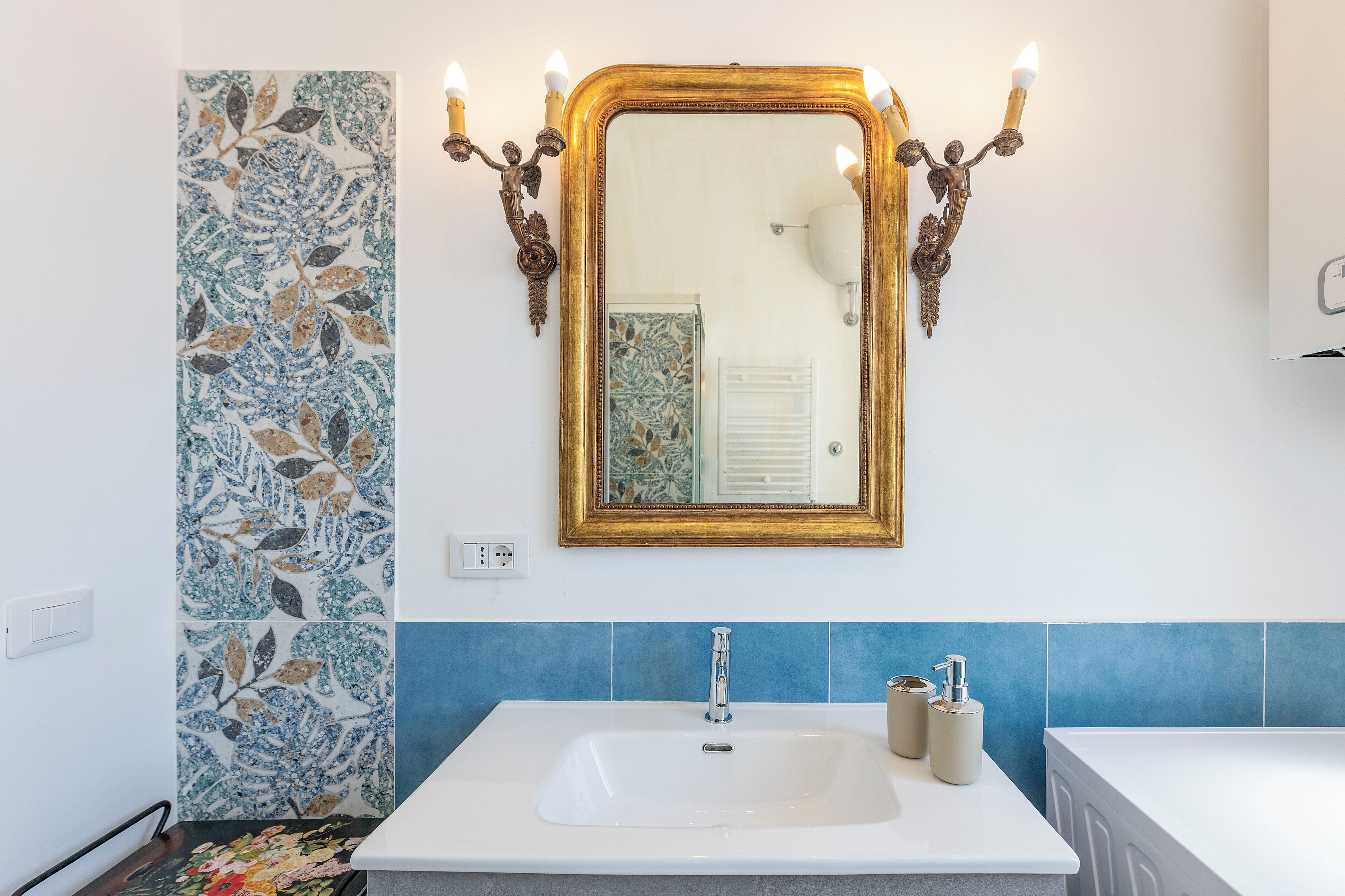 Un baño con un lavabo blanco, azulejos azules, un espejo con marco dorado y una lámpara de pared.