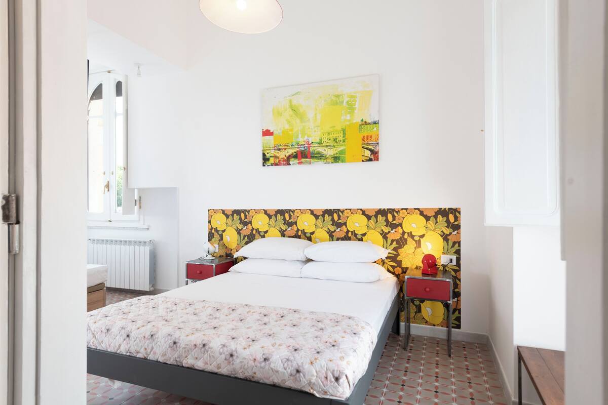 A bedroom with a bed, a nightstand, a red lamp, and a floral-patterned headboard.
