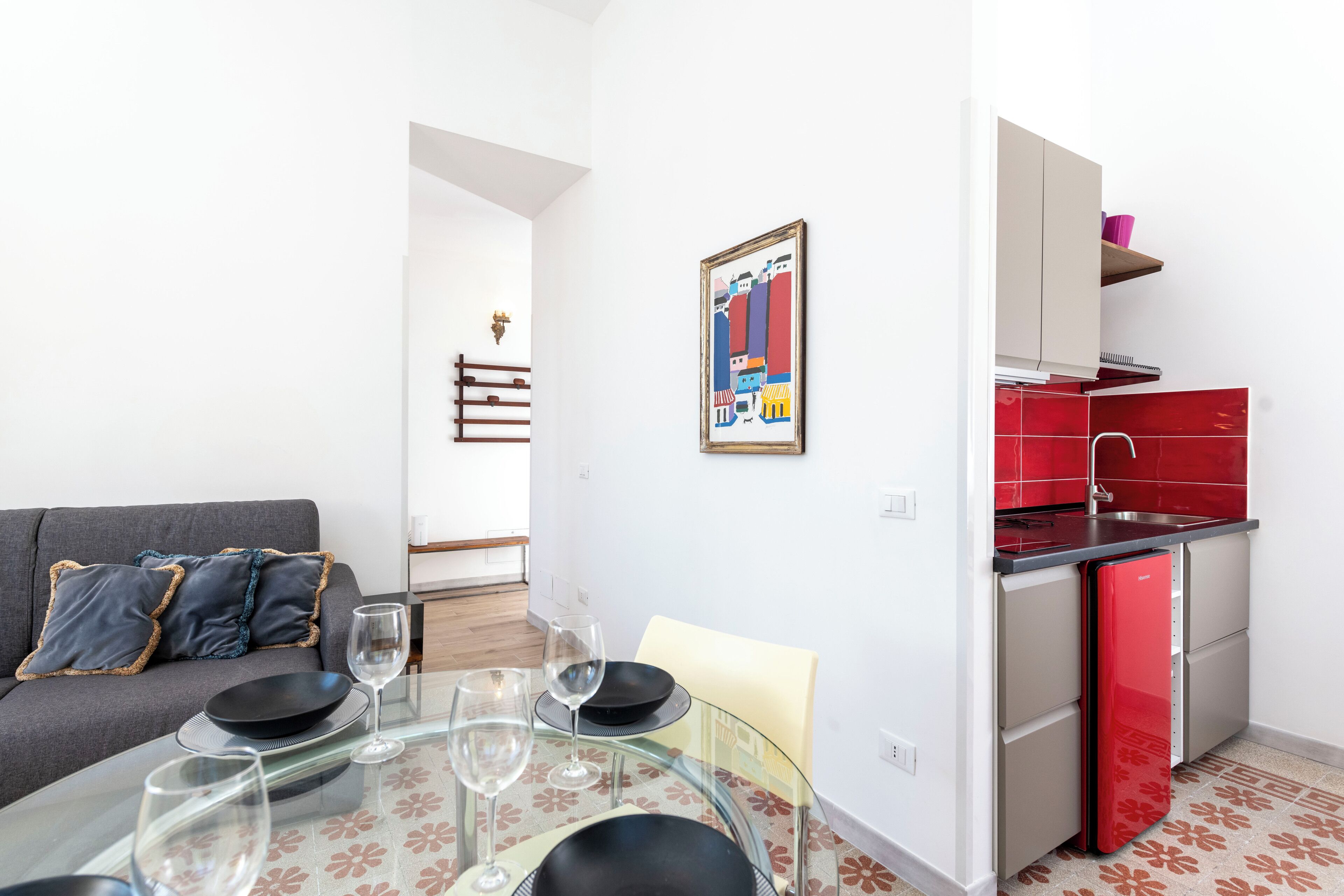 A compact living space with a dining table set for two, a sofa with decorative pillows, a kitchen with red appliances, and a framed artwork on the wall.