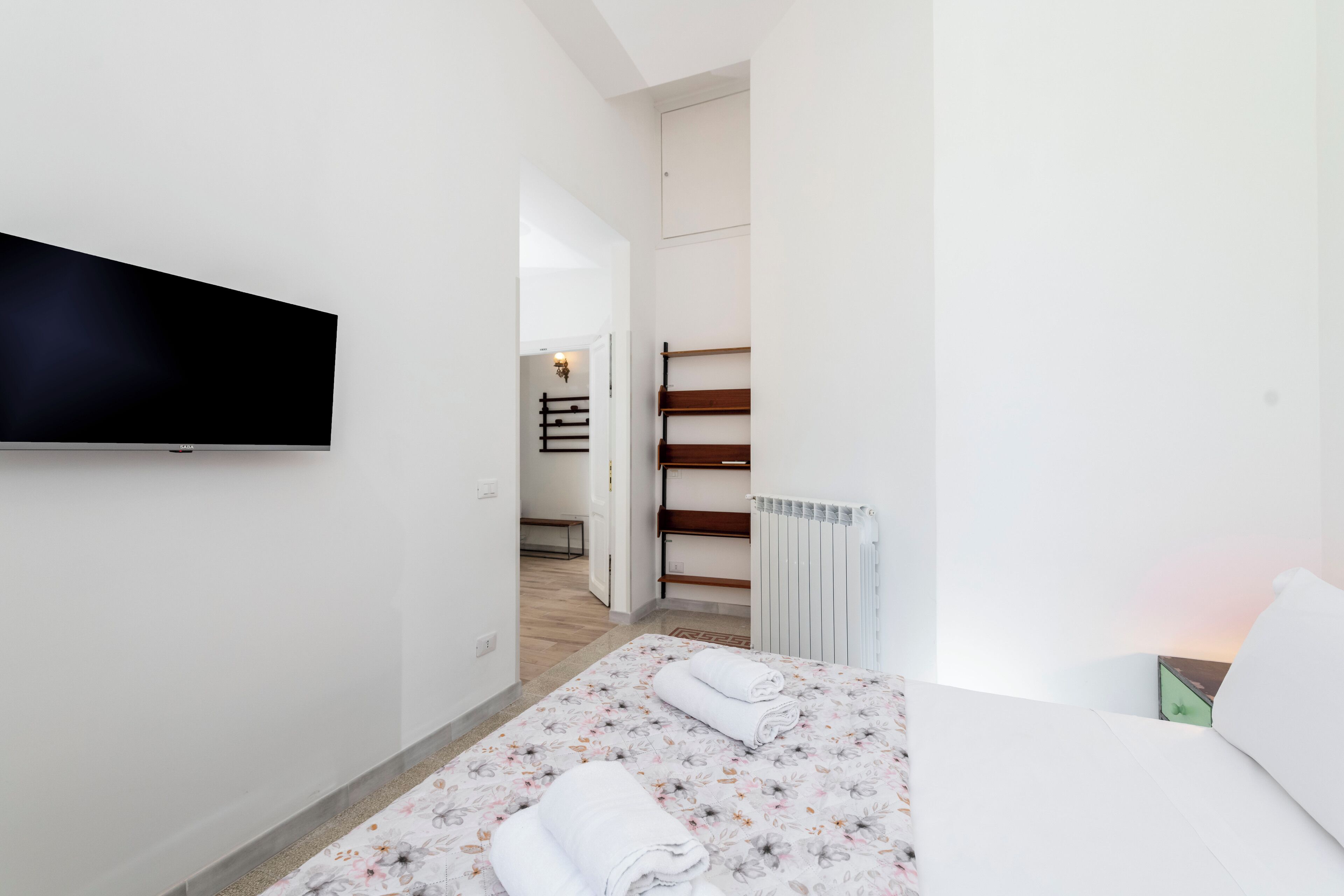 Una habitació d'hotel moderna amb televisió de pantalla plana penjada a la paret, un llit amb roba de llit floral, un radiador i vista al passadís.