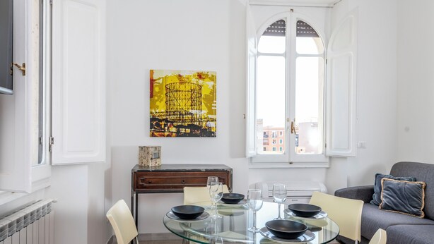 A modern dining area with a glass table, white chairs, and black bowls. A painting hangs on the wall, and a sofa is visible in the background. - Trastevere novecento (Rome)