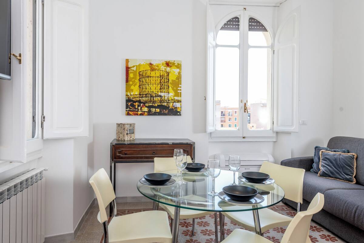 A modern dining area with a glass table, white chairs, and black bowls. A painting hangs on the wall, and a sofa is visible in the background.