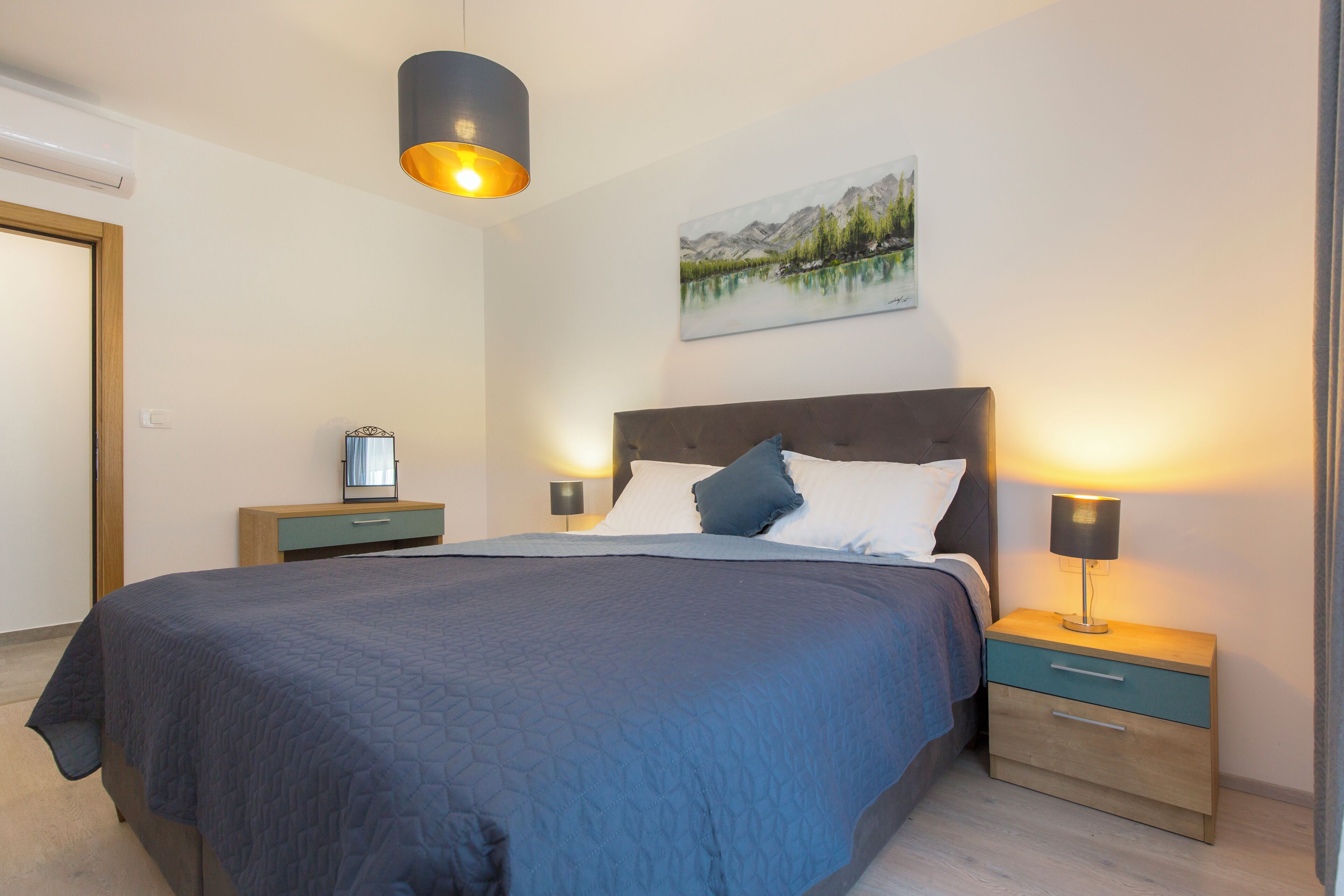 A neatly arranged bedroom with a large bed, two bedside tables, a nightstand, a wall-mounted lamp, and a painting of a mountainous landscape.