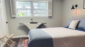 Ein Schlafzimmer mit einem Bett, einem Nachttisch, einem Fenster mit Rollläden, einer Wandlampe und einem kleinen Tisch mit einer Dekoration.