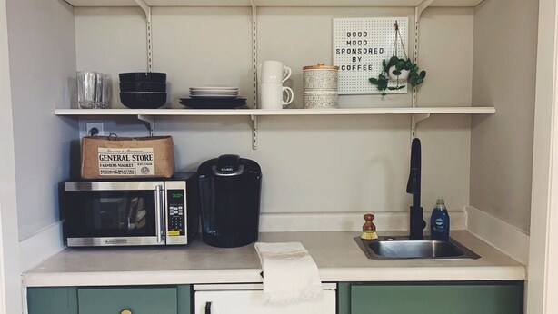 Eine kompakte Küche mit Mikrowelle, Kaffeemaschine und kleinem Kühlschrank. Darüber befinden sich Regale mit verschiedenen Küchenutensilien und ein Schild.