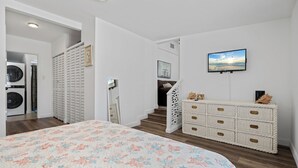 A bedroom with a floral bedspread, a white dresser with drawers, a television mounted on the wall, and a staircase leading to another room.