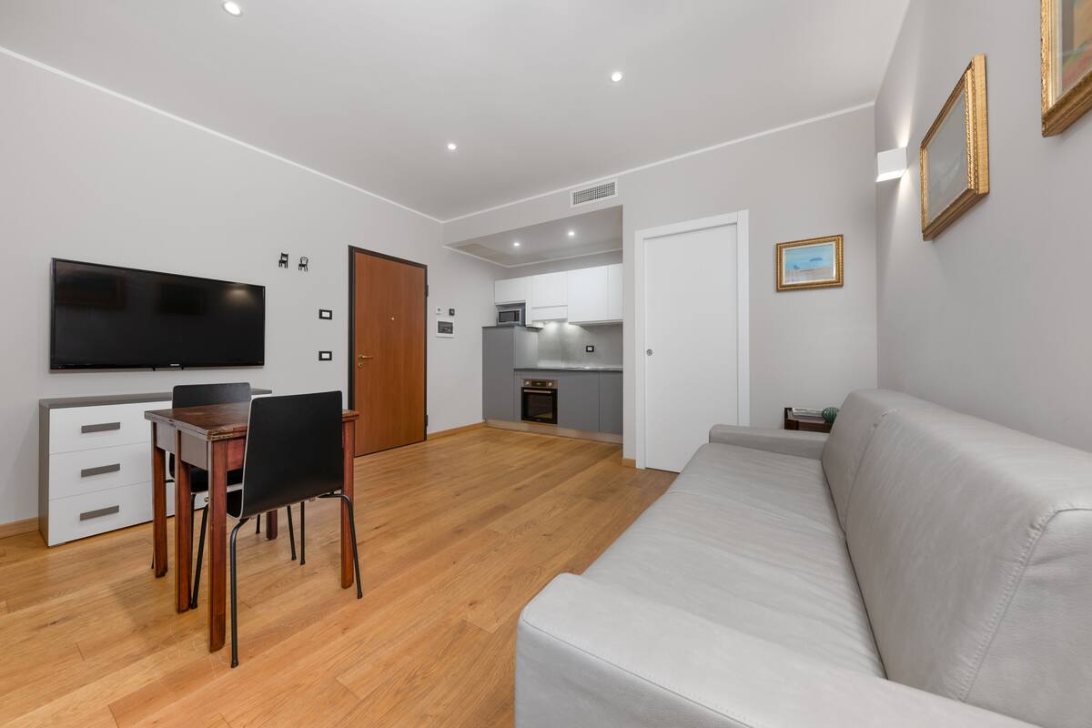 A modern living room with a flat-screen TV, a dining table with chairs, a sofa, and a kitchen area in the background.