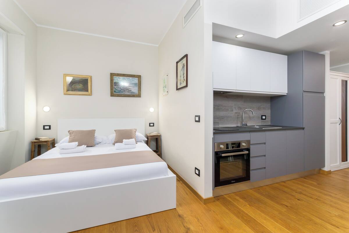 A modern bedroom with a large bed, a small bedside table, and a nightstand. Adjacent to the bedroom is a compact kitchen with a sink, stove, and cabinets.