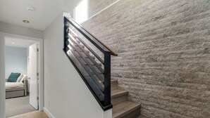 Una escalera con escalones de madera y barandillas negras, que lleva a una habitación con cama y vistas a un muro de piedra.