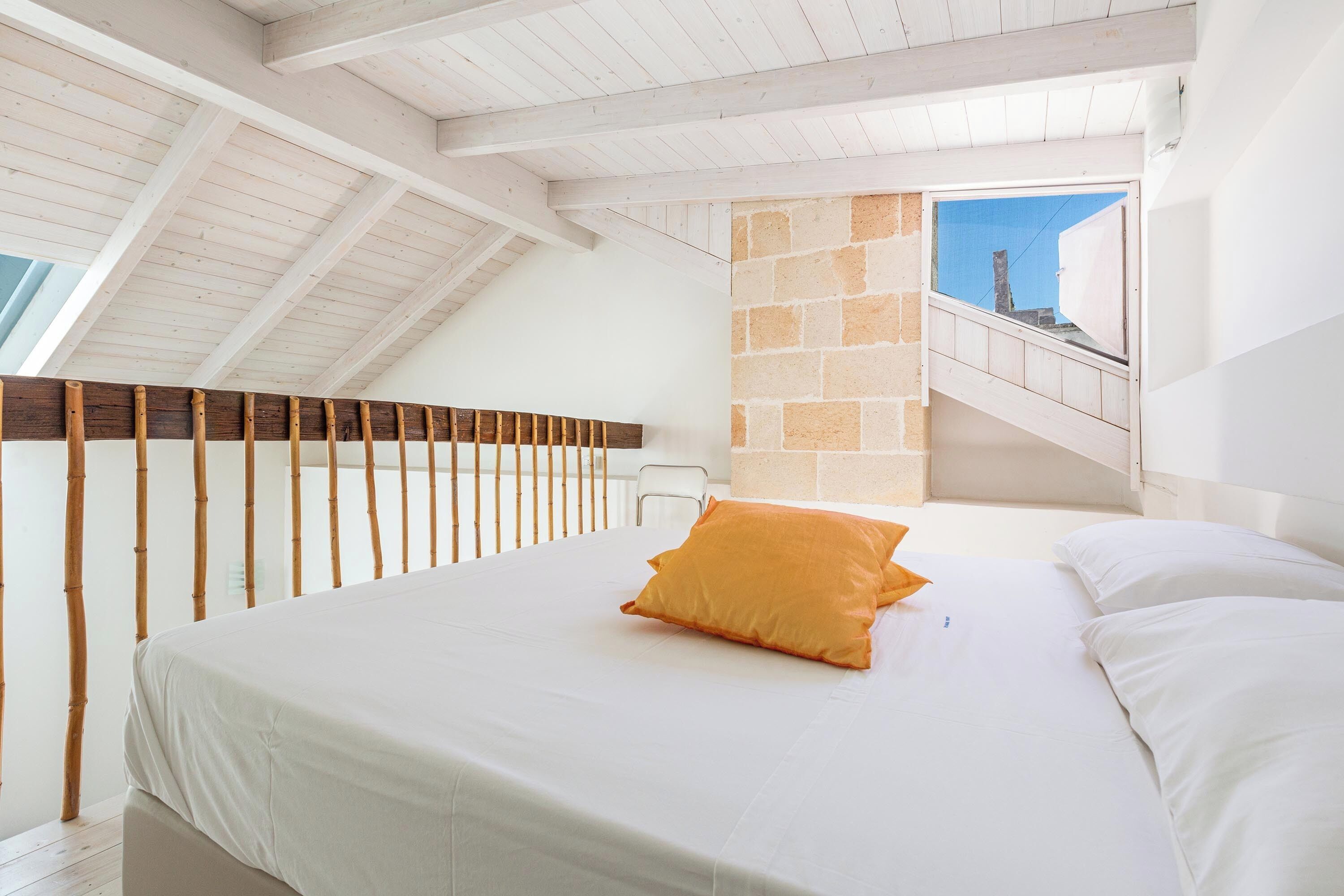 A bedroom with a sloped ceiling, a bed with white linens and an orange pillow, a wooden railing, and a stone wall with a window.