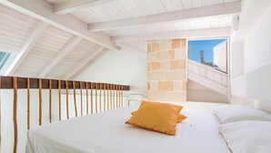 Ein Schlafzimmer mit schrägen Wänden, einem Bett mit weißen Leinen und einem orangefarbenen Kissen, einem Holzlaufgeländer und einer Steinwand mit einem Fenster.