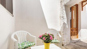 Ein kleiner Sitzbereich im Freien mit zwei weißen Plastikstühlen und einem Tisch, einem gelben Topf mit rosa Blumen und einer Holztür im Hintergrund.