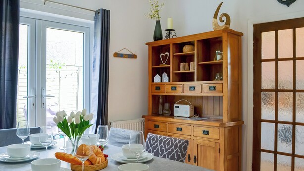 Un comedor con una mesa preparada para dos personas, un mueble de madera con adornos, y una vista a través de una puerta con vidrio esmerilado.